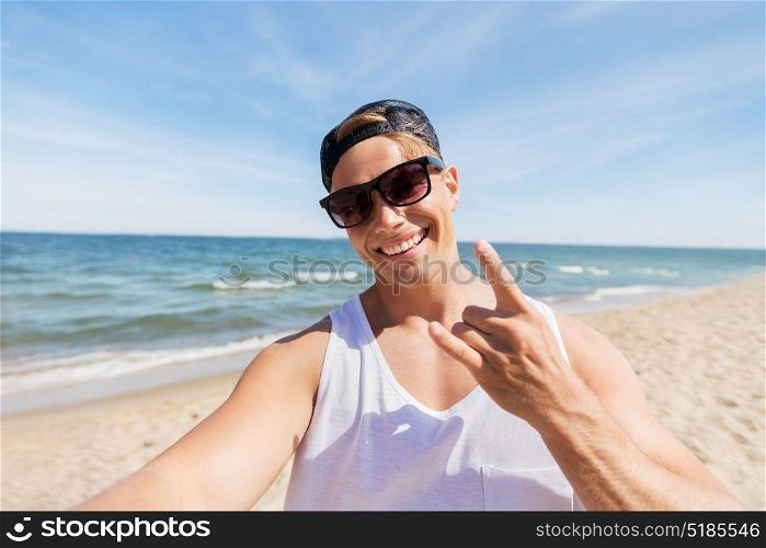 summer holidays, gesture and people concept - happy smiling young man in sunglasses and hat taking selfie and showing rock sign on beach. man in sunglasses taking selfie on summer beach