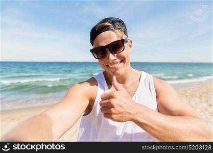 summer holidays, gesture and people concept - happy smiling young man in sunglasses and hat taking selfie and showing thumbs up on beach. man in sunglasses taking selfie on summer beach