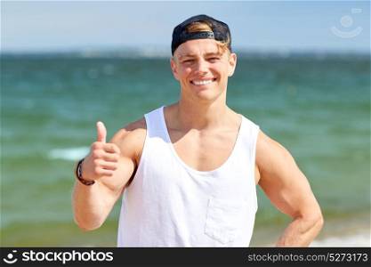 summer holidays, gesture and people concept - happy smiling young man wearing cap showing thumbs up on beach. smiling man showing thumbs up on summer beach