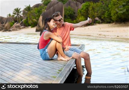 summer holidays, love and people concept - happy teenage couple taking selfie on smartphone and sitting on berth over exotic seychelles beach background. happy teenage couple taking selfie on smartphone