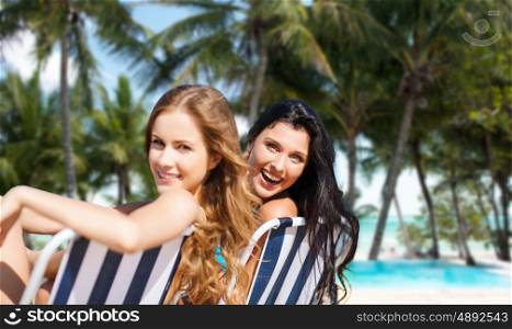 summer holidays, people, leisure, vacation and travel concept - happy women sunbathing in chairs over exotic tropical beach with palm trees and pool background