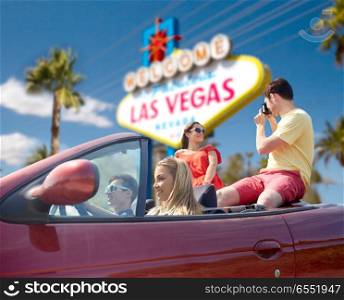 summer holidays, road trip and travel concept - happy friends driving in convertible car and taking picture by film camera over welcome to fabulous las vegas sign background. friends with camera driving in car at las vegas. friends with camera driving in car at las vegas