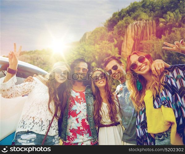 summer holidays, road trip, travel and people concept - smiling young hippie friends at minivan car showing peace hand sign over island background. hippie friends at minivan car showing peace sign