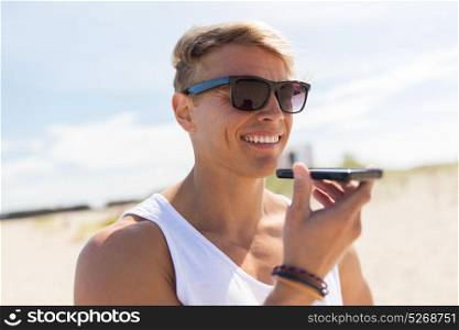 summer holidays, technology and people concept - happy smiling young man in sunglasses using voice command recorder on smartphone on beach. smiling man calling on smartphone at summer beach