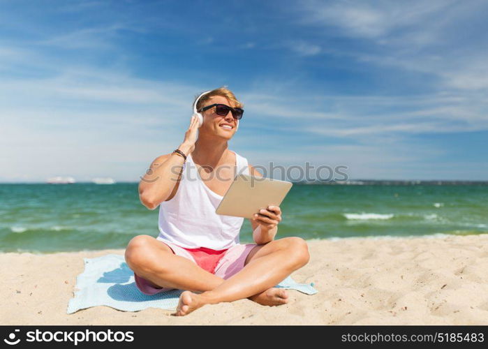 summer holidays, technology and people concept - happy smiling young man with tablet pc computer and headphones listening to music and sunbathing on beach towel. happy man with tablet pc and headphones on beach
