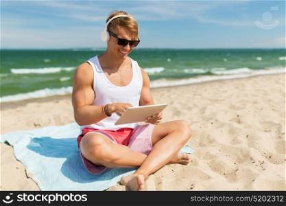 summer holidays, technology and people concept - happy smiling young man with tablet pc computer and headphones listening to music and sunbathing on beach towel. happy man with tablet pc and headphones on beach