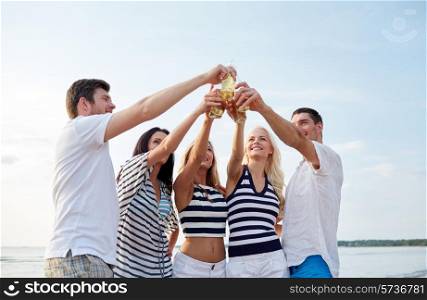 summer, holidays, tourism, drinks and people concept - group of smiling friends clinking bottles of beer or cider on beach