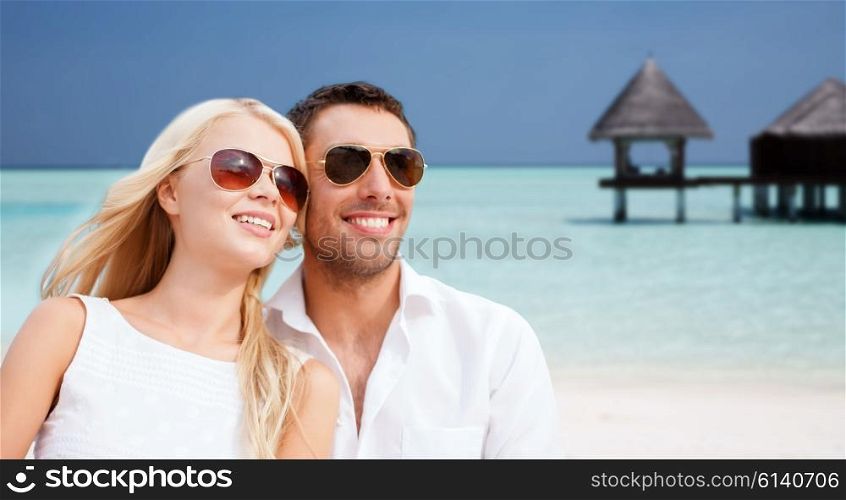 summer holidays, tourism, vacation, travel and dating concept - happy couple in shades at sea side over beach with bungalow background