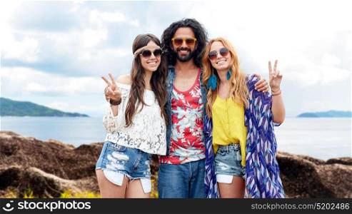 summer holidays, travel and people concept - smiling young hippie friends in sunglasses showing peace hand sign over sea and island background. happy hippie friends showing peace on summer beach