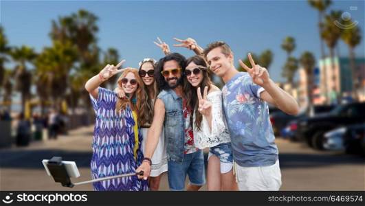 summer holidays, travel and technology concept - smiling young hippie friends taking picture by smartphone on selfie stick and showing peace gesture over venice beach in los angeles background. hippie friends with smartphone on selfie stick