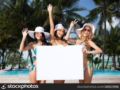 summer holidays, travel, people, advertisement and vacation concept - happy young women in bikinis holding white board over exotic tropical beach with palm trees and pool background