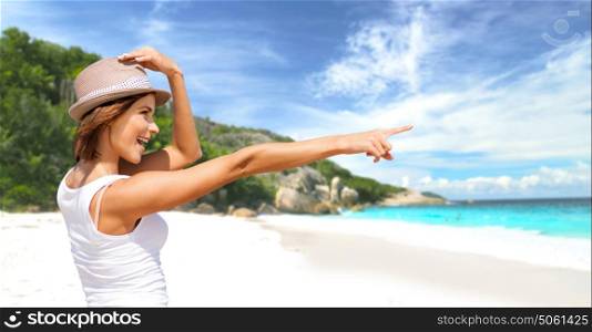 summer holidays, travel, people and vacation concept - happy smiling young woman in hat pointing finger over exotic tropical beach with palm trees and sea shore background. happy young woman in hat on summer beach