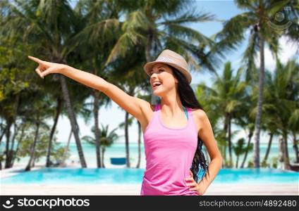summer holidays, travel, people and vacation concept - happy young woman in hat pointing finger over exotic tropical beach with palm trees and pool background