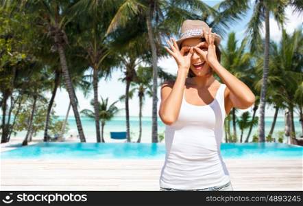 summer holidays, travel, people and vacation concept - happy young woman in hat making finger glasses over exotic tropical beach with palm trees and pool background