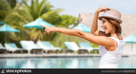 summer holidays, travel, people and vacation concept - happy young woman in hat pointing finger over pool with palm trees and sunbeds background. happy young woman in hat on summer beach