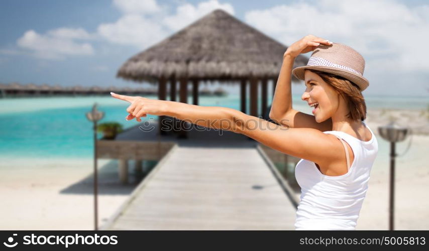 summer holidays, travel, people and vacation concept - happy young woman in hat pointing finger over exotic tropical beach with bungalow and sea shore background. happy young woman in hat on summer beach