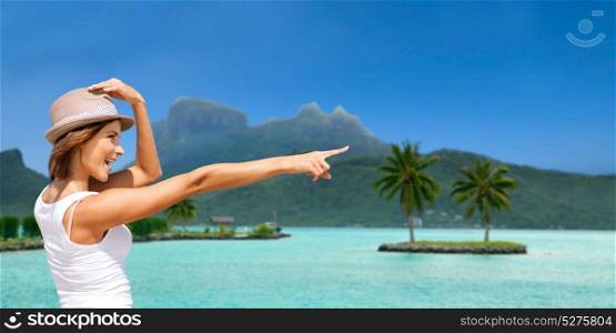 summer holidays, travel, people and vacation concept - happy young woman in hat pointing finger at touristic resort over exotic bora bora island beach background. happy young woman in hat on bora bora beach