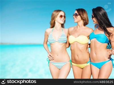 summer holidays, travel, people and vacation concept - happy young women in bikinis and shades over blue sky and sea background