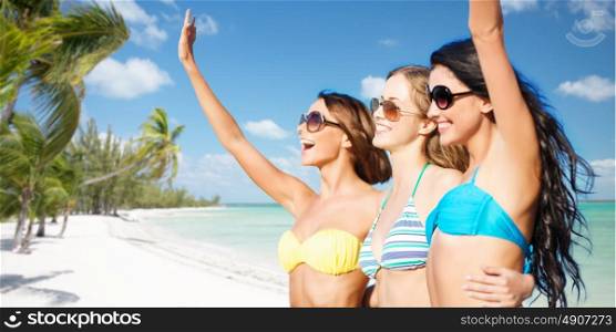 summer holidays, travel, people and vacation concept - happy young women in bikinis and shades hugging and waving hands over exotic tropical beach with palm trees and sea shore background. happy young women in bikinis on summer beach