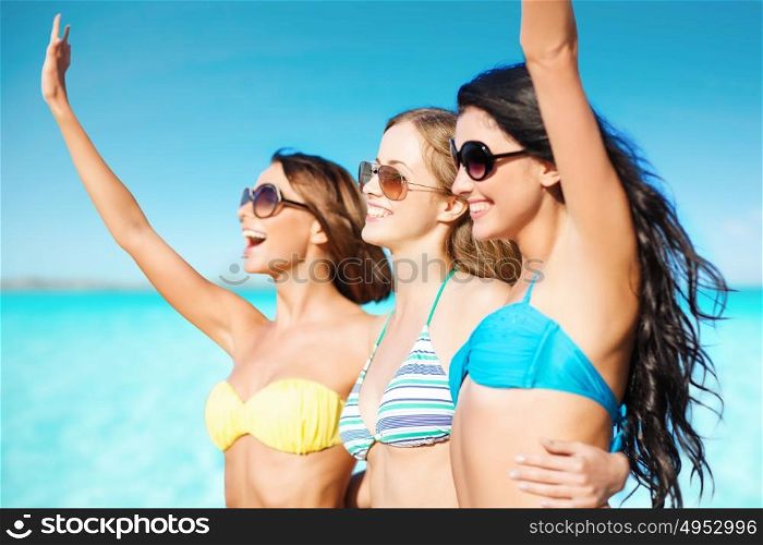 summer holidays, travel, people and vacation concept - happy young women in bikinis and shades hugging and waving hands on beach over sea and blue sky background. happy young women in bikinis on summer beach
