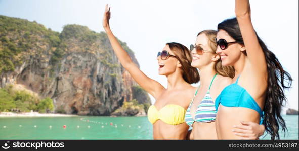 summer holidays, travel, people and vacation concept - happy young women in bikinis and shades hugging and waving hands over bali beach and rock background. happy young women in bikinis on bali beach