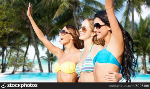 summer holidays, travel, people and vacation concept - happy young women in bikinis and shades hugging and waving hands over exotic tropical beach with palm trees and pool background. happy young women in bikinis on summer beach