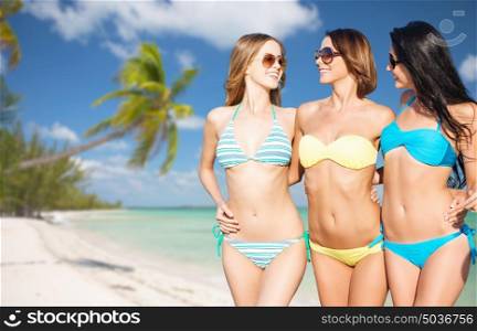 summer holidays, travel, people and vacation concept - happy young women in bikinis and shades over exotic tropical beach with palm trees and sea shore background. happy young women in bikinis on summer beach