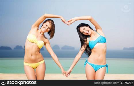 summer holidays, travel, people, love and vacation concept - happy young women in bikinis making heart shape with hands over infinity edge pool background
