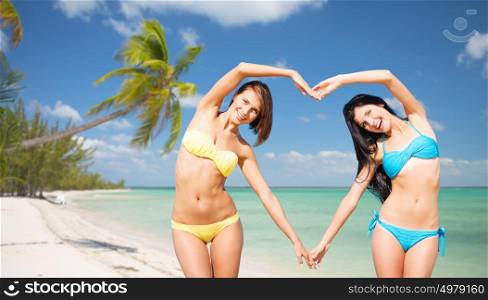 summer holidays, travel, people, love and vacation concept - happy young women in bikinis making heart shape with hands over exotic tropical beach with palm trees and sea shore background. happy women making heart shape on summer beach