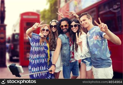 summer holidays, travel, technology and people concept - smiling young hippie friends taking picture by smartphone on selfie stick and showing peace gesture over london city street background
