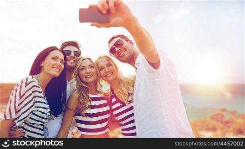 summer holidays, travel, tourism, technology and people concept - group of smiling friends with smartphone photographing and taking selfie over exotic beach background