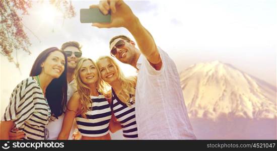 summer holidays, travel, tourism, technology and people concept - group of smiling friends with smartphone photographing and taking selfie over fujiyama mountain in japan background