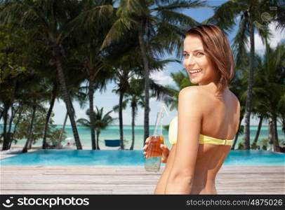 summer holidays, vacation and people concept - happy smiling young woman in bikini with bottle of drink over exotic tropical beach with palm trees and pool background