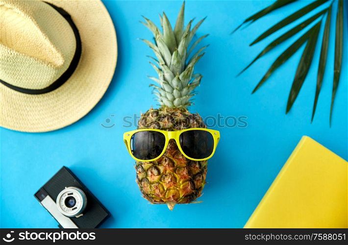 summer holidays, vacation and travel concept - close up of pineapple in yellow sunglasses, hat, camera and palm leaf on blue background. pineapple in sunglasses, hat, camera and palm leaf