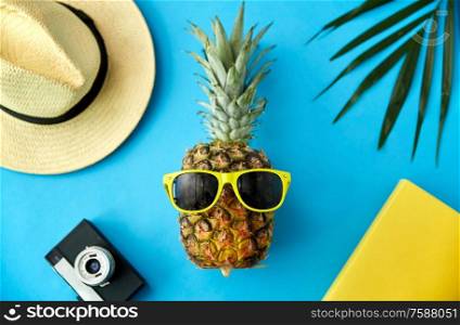summer holidays, vacation and travel concept - close up of pineapple in yellow sunglasses, hat, camera and palm leaf on blue background. pineapple in sunglasses, hat, camera and palm leaf