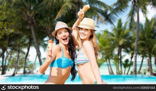 summer holidays, vacation, food, travel and people concept - smiling young women eating ice cream over exotic tropical beach with palm trees and pool background. smiling women eating ice cream on beach