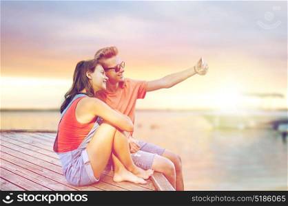 summer holidays, vacation, love and people concept - happy teenage couple taking selfie on smartphone sitting on berth over sea background. happy teenage couple taking selfie on smartphone