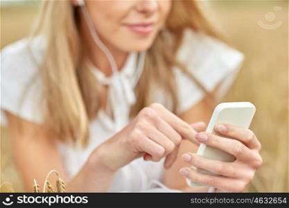 summer holidays, vacation, technology and people concept - close up of smiling young woman in white dress with smartphone and earphones listening to music on cereal field