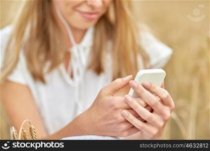 summer holidays, vacation, technology and people concept - close up of smiling young woman in white dress with smartphone and earphones listening to music on cereal field