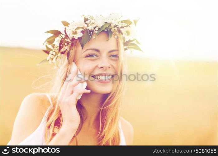 summer holidays, vacation, technology and people concept - close up of smiling young woman in wreath of flowers calling on smartphone at countryside. happy young woman calling on smartphone at country