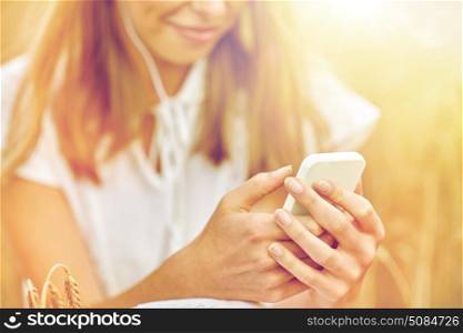summer holidays, vacation, technology and people concept - close up of smiling young woman in white dress with smartphone and earphones listening to music on cereal field. close up of woman with smartphone and earphones. close up of woman with smartphone and earphones