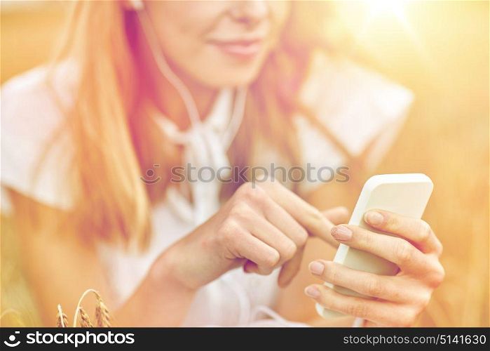 summer holidays, vacation, technology and people concept - close up of smiling young woman in white dress with smartphone and earphones listening to music on cereal field. close up of woman with smartphone and earphones