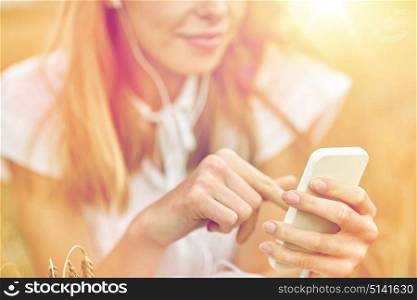 summer holidays, vacation, technology and people concept - close up of smiling young woman in white dress with smartphone and earphones listening to music on cereal field. close up of woman with smartphone and earphones