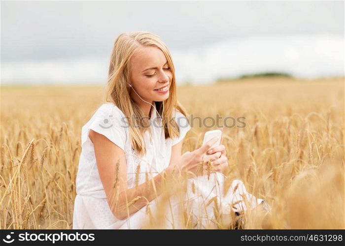 summer holidays, vacation, technology and people concept - smiling young woman in white dress with smartphone and earphones listening to music on cereal field