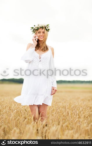 summer holidays, vacation, technology and people concept - smiling young woman in wreath of flowers calling on smartphone on cereal field