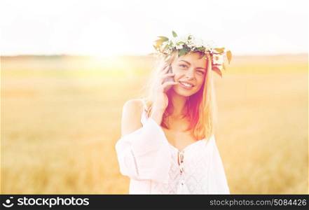 summer holidays, vacation, technology and people concept - smiling young woman in wreath of flowers calling on smartphone on cereal field. happy young woman calling on smartphone at country. happy young woman calling on smartphone at country