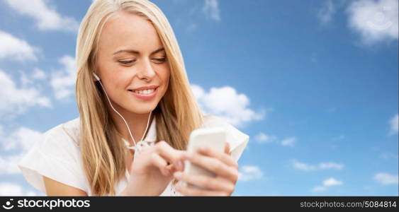 summer holidays, vacation, technology and people concept - smiling young woman in white dress with smartphone and earphones listening to music over blue sky and clouds background. happy woman with smartphone and earphones over sky. happy woman with smartphone and earphones over sky