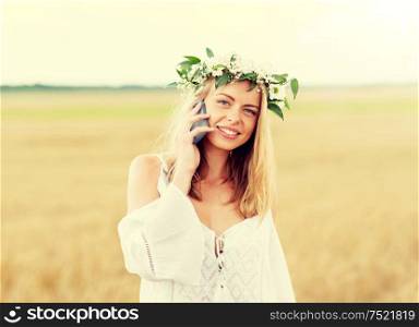 summer holidays, vacation, technology and people concept - smiling young woman in wreath of flowers calling on smartphone on cereal field. happy young woman calling on smartphone at country