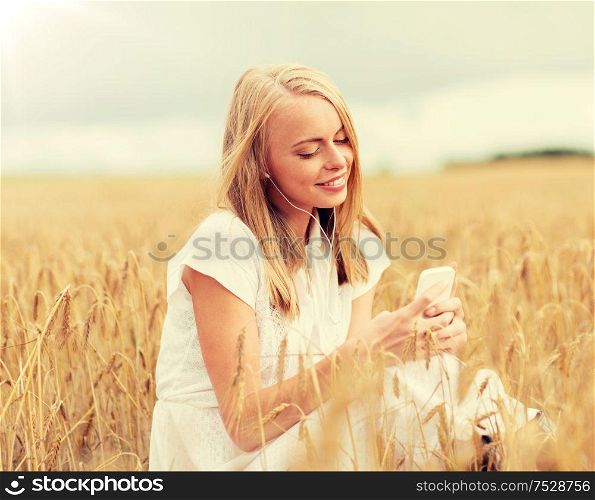 summer holidays, vacation, technology and people concept - smiling young woman in white dress with smartphone and earphones listening to music on cereal field. happy woman with smartphone and earphones
