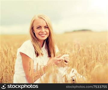summer holidays, vacation, technology and people concept - smiling young woman in white dress with smartphone and earphones listening to music on cereal field. happy woman with smartphone and earphones